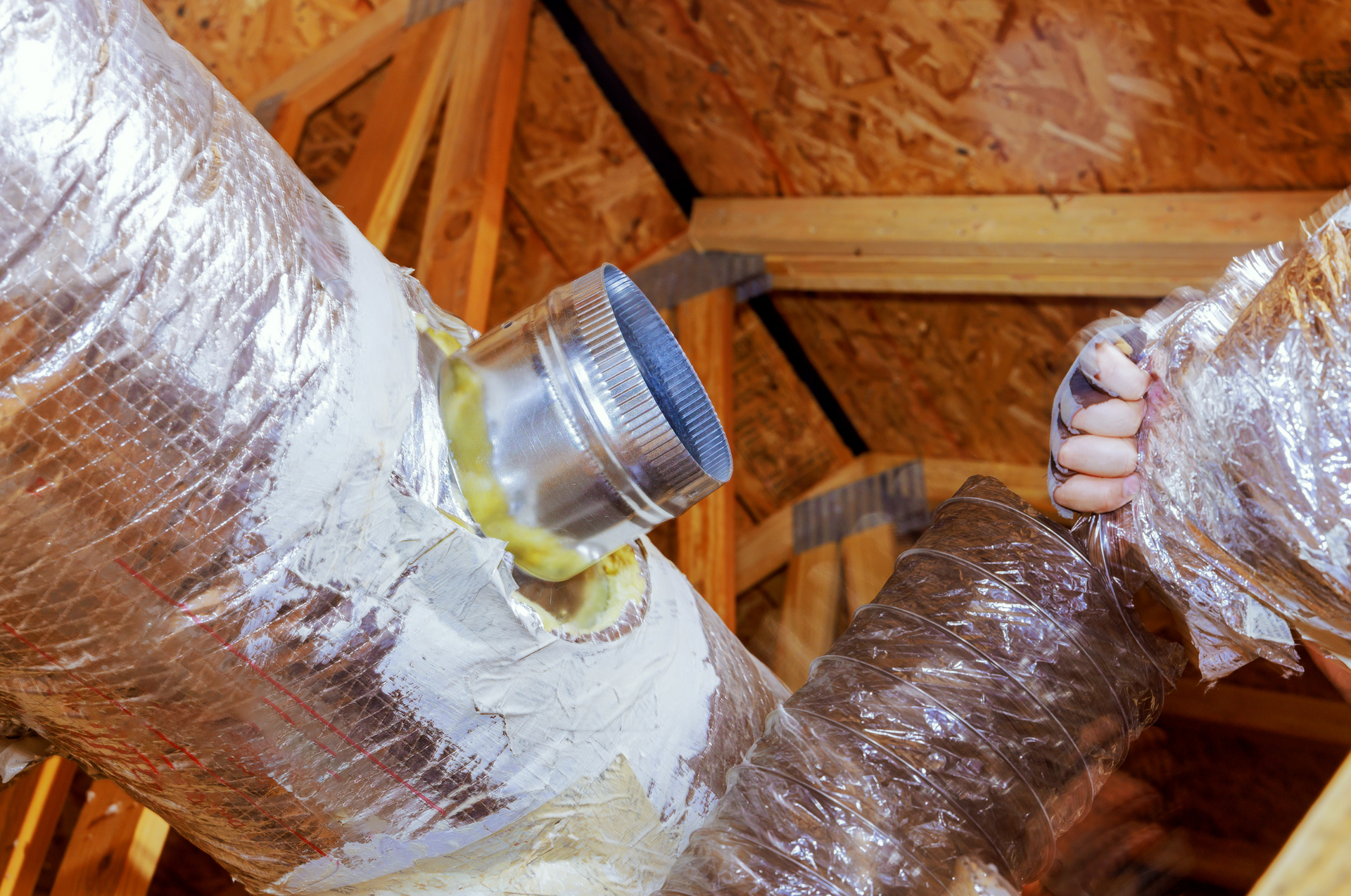 HVAC ductwork connection in an attic, featuring foil-wrapped flexible ducts, a metal fitting, and exposed wood framing.