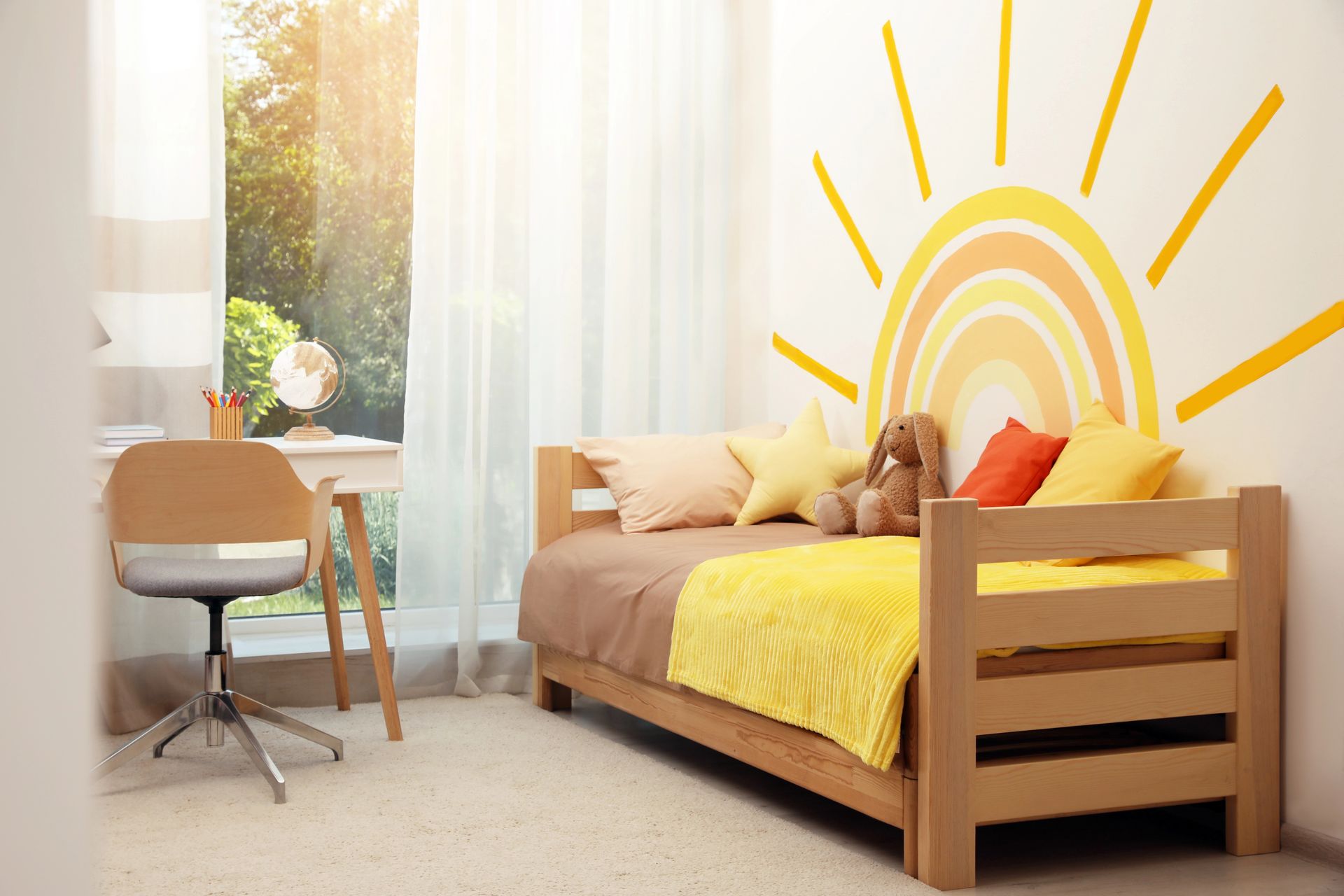 A cozy child's bedroom with a wooden bed, yellow sun wall mural, desk, and chair bathed in natural light.