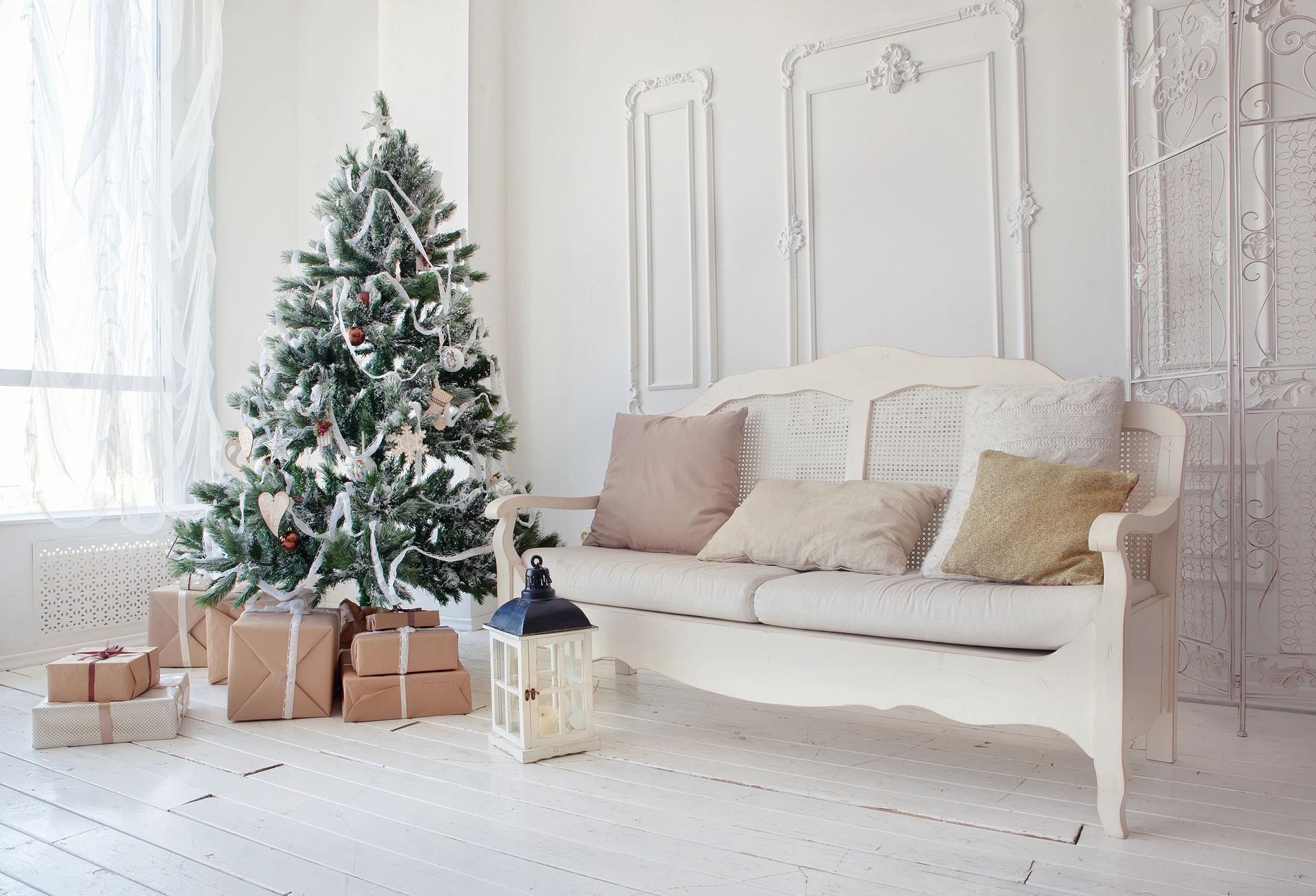 Christmas tree with wrapped gifts, a white couch with pillows, and a lantern in a bright room.