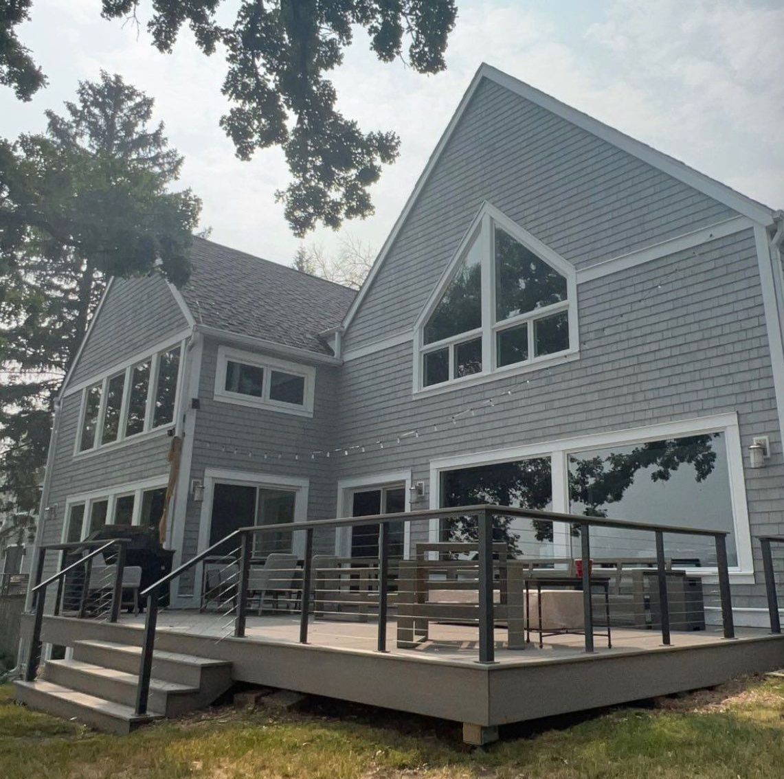 Gray house with a deck overlooking a body of water, featuring multiple windows and a gabled roof.