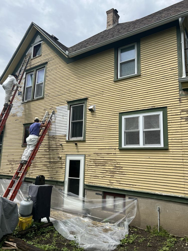 A man on a ladder is scraping paint off the side of a house.