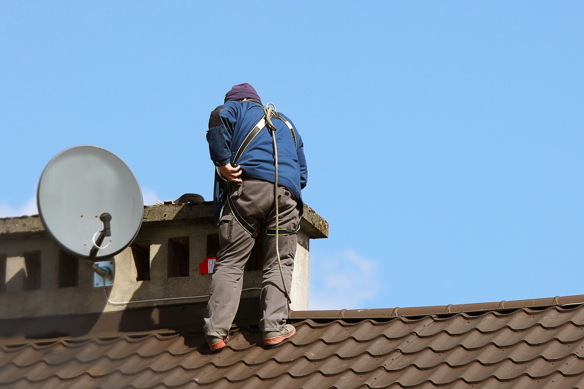 Person wearing safety harness on rooftop, near a chimney and satellite dish. Person wearing safety harness on rooftop, near a chimney and satellite dish.
