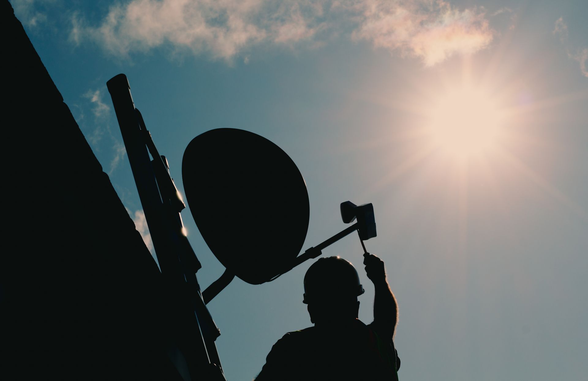 Silhouette of a person adjusting a satellite dish on a building with a sunny sky in the background.