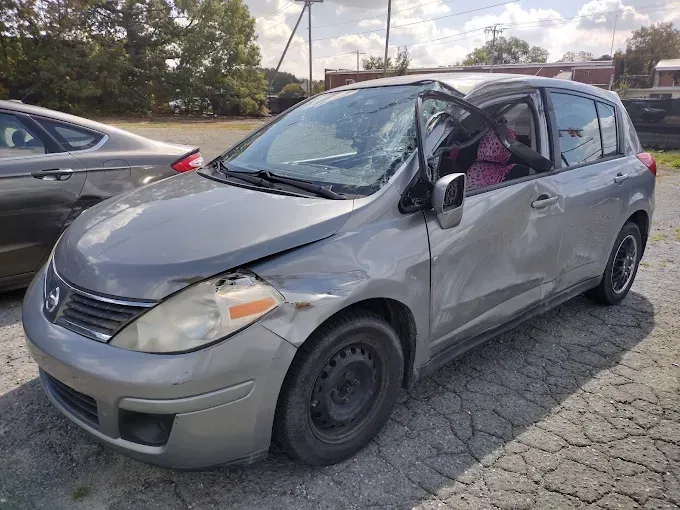 Damaged silver Nissan Versa hatchback with front and side impact damage, parked outdoors.
