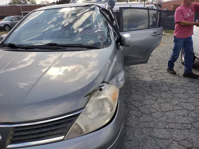 Damaged silver Nissan Versa with smashed windshield and dented front, next to a person.