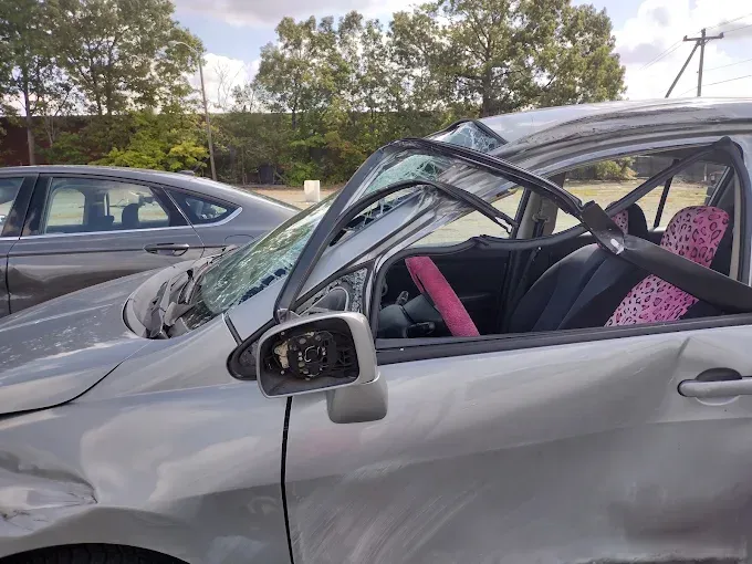Silver car damaged in accident, side view with broken windshield, pink details inside.
