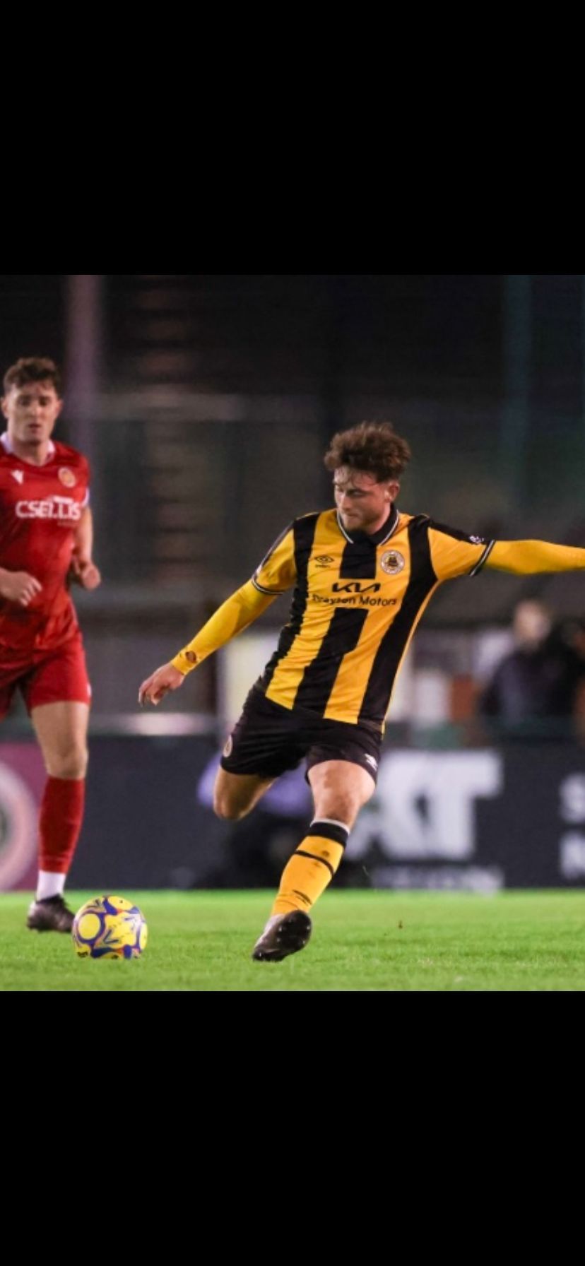 Soccer player kicking a ball, wearing a black and gold striped jersey, with another player nearby.
