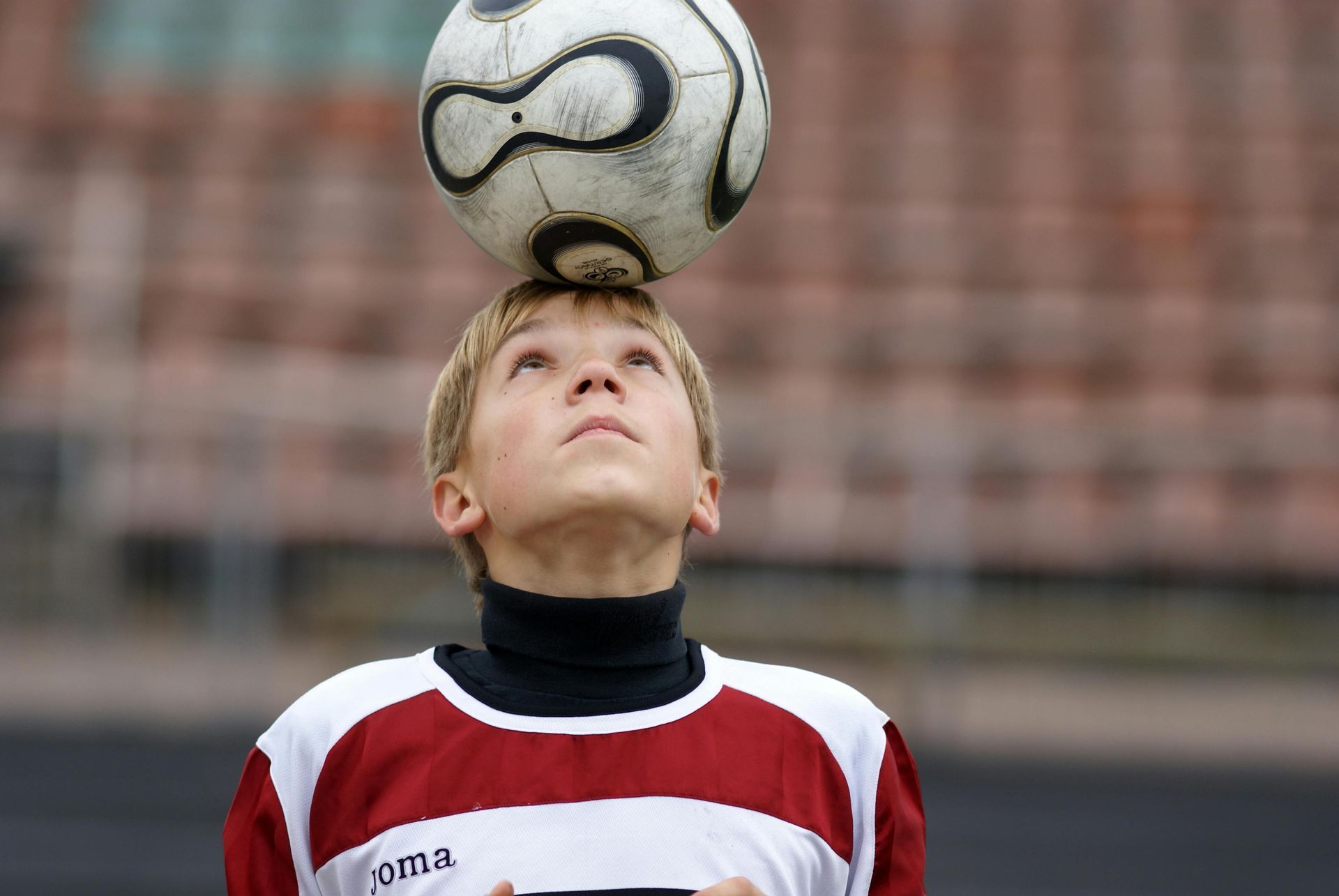 Boy in soccer jersey balancing ball on head in stadium.
