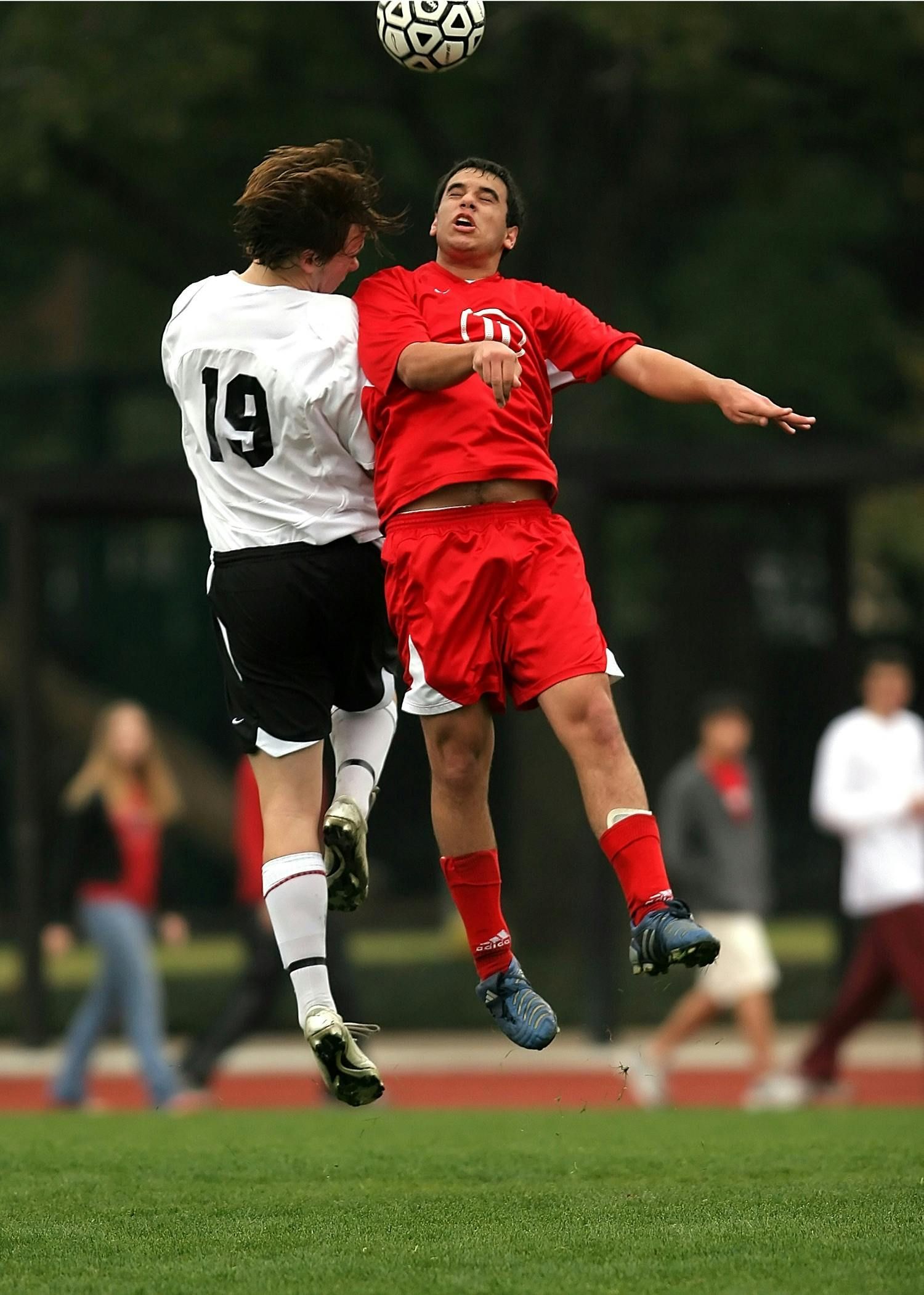 Two soccer players jump for a high ball, one in red and one in white, on a field.