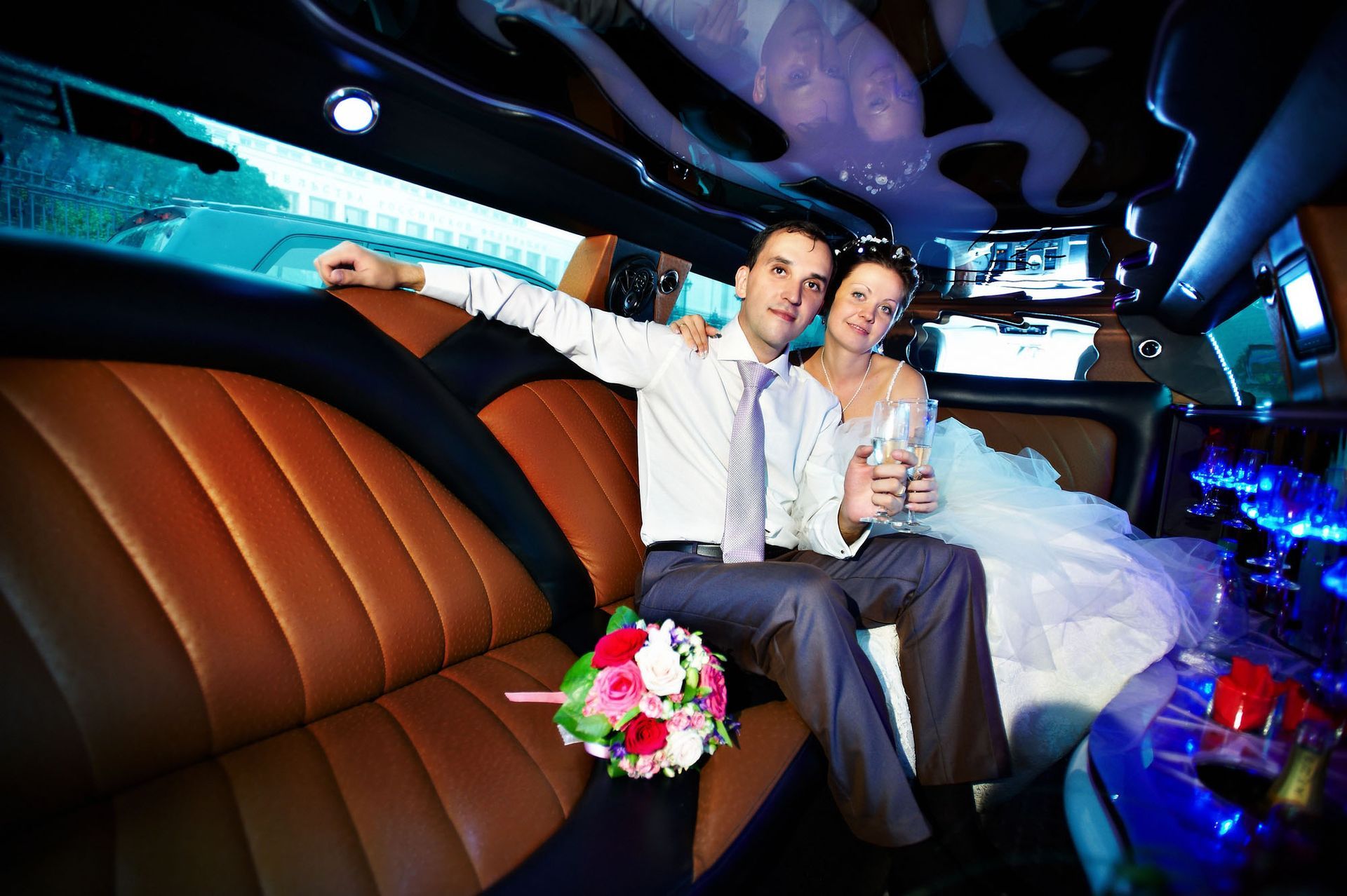 Newlyweds in a limo, man with arm around woman in a white dress, holding a bouquet, brown leather seats.