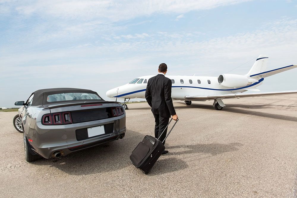 Man in suit with suitcase walks towards private jet and convertible car on tarmac.