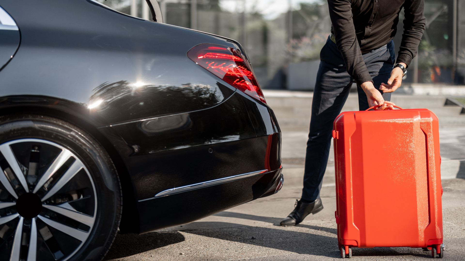 Person loading a bright red suitcase into the trunk of a black car. Outdoor setting, sunny day.