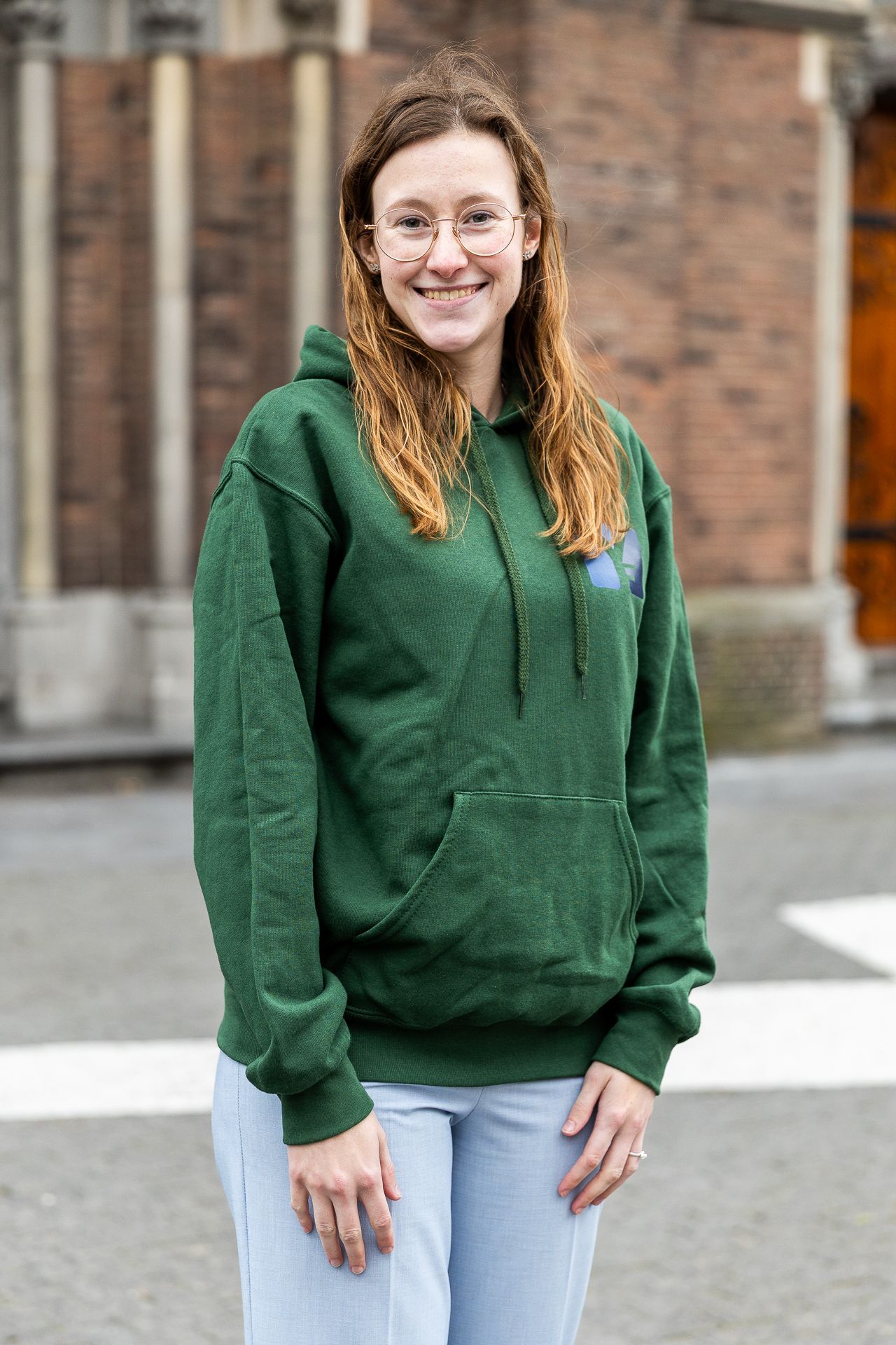 Een vrouw in een groene hoodie en met een bril staat lachend voor een bakstenen gebouw.
