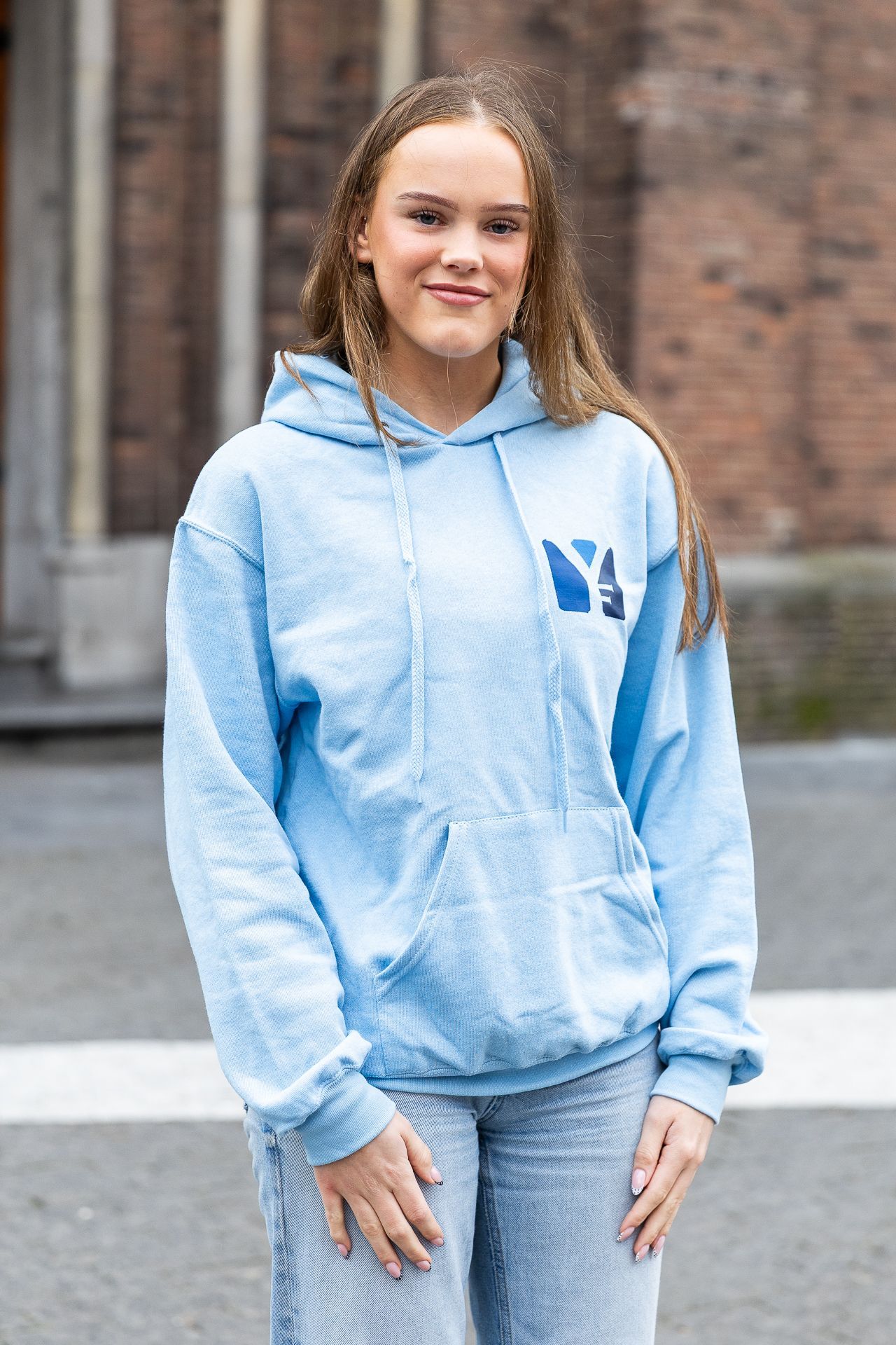 Een persoon in een lichtblauwe hoodie en spijkerbroek staat lachend voor een bakstenen gebouw.