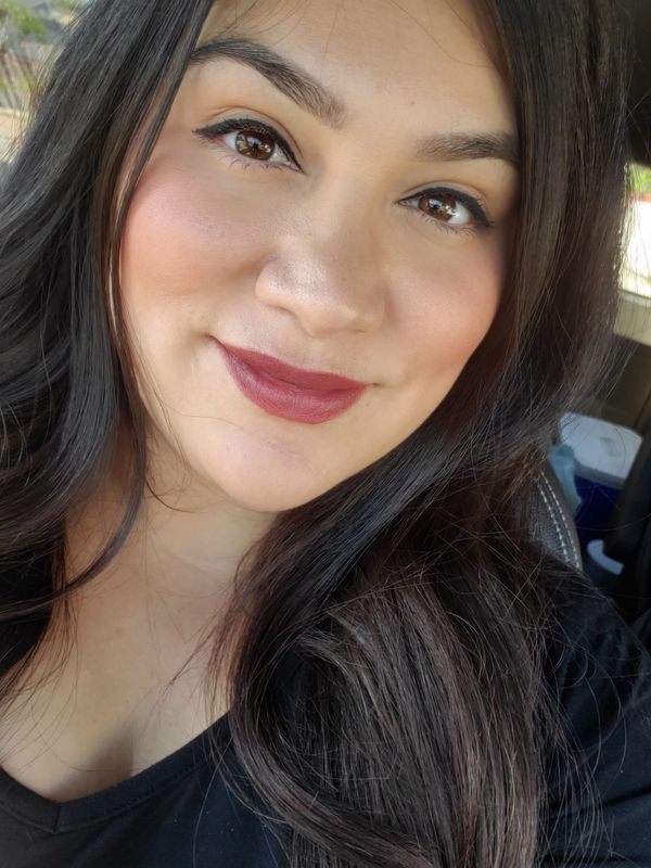 Woman with dark hair, wearing makeup, smiling, with burgundy lipstick.