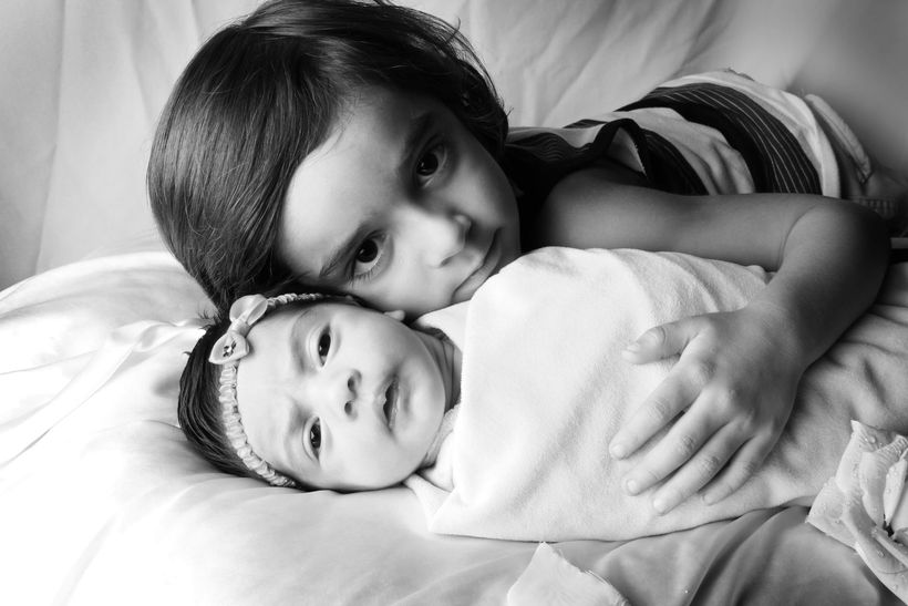 Older child lovingly embraces a swaddled infant on a bed, black and white photo.