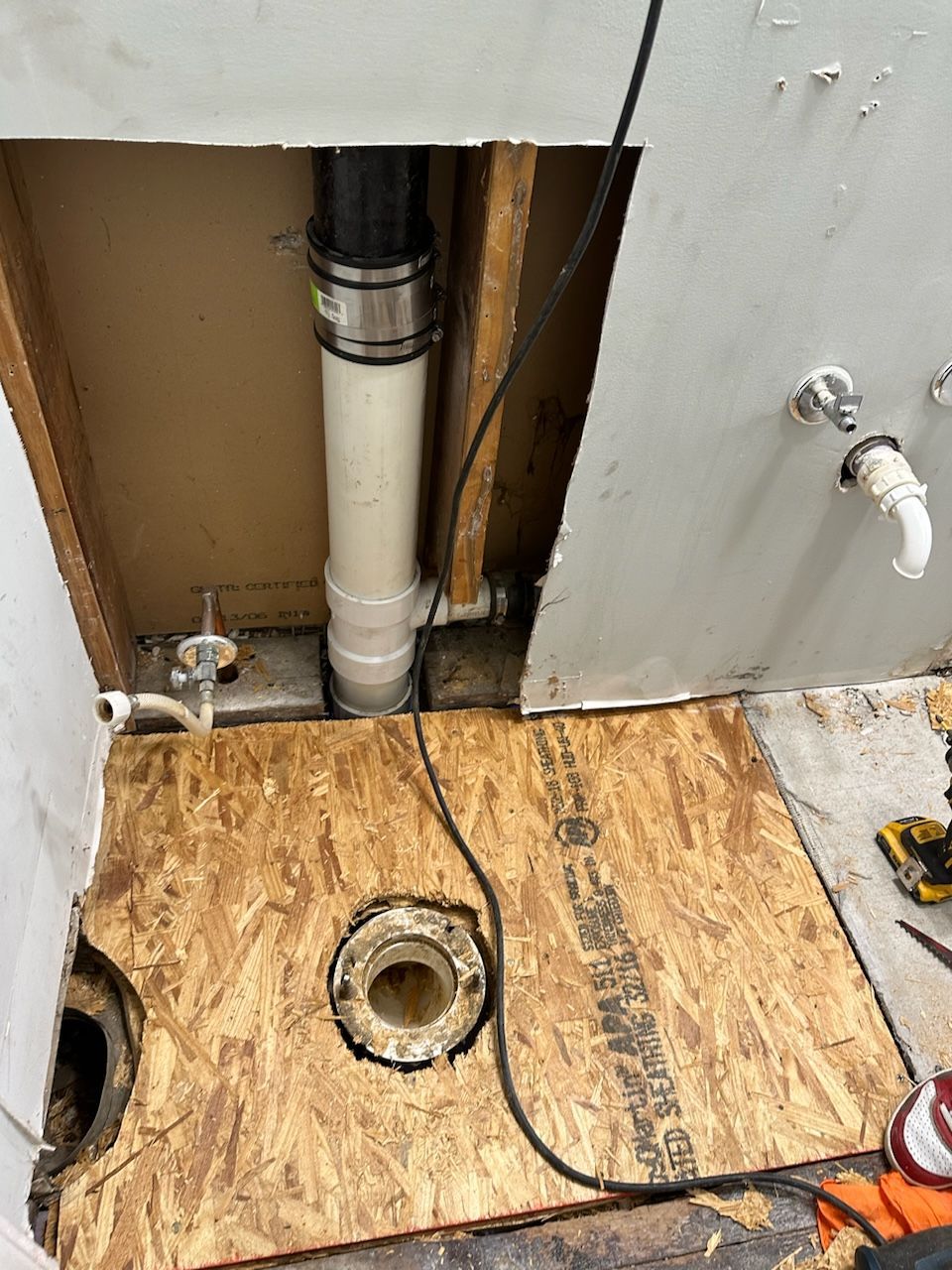 Bathroom under construction, plumbing visible. Pipes, exposed floor, wood, and a sink drain are shown.