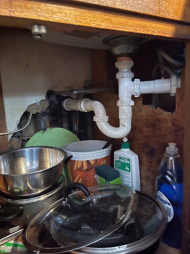 Under-sink view of white plumbing pipes, with food items and cleaning supplies in a cabinet.