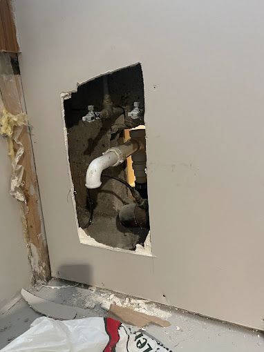 Hole in wall exposing plumbing pipes. White wall, grey floor.
