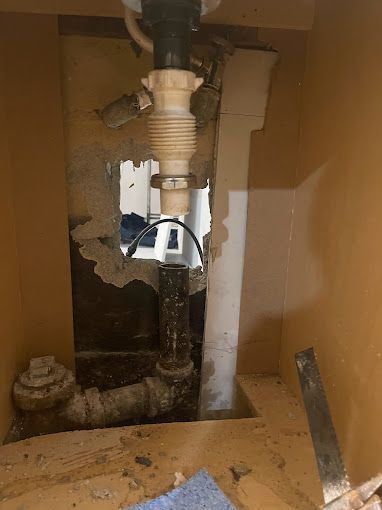 Under-sink plumbing with a flexible drain pipe and exposed wall opening. Brown cabinet interior.