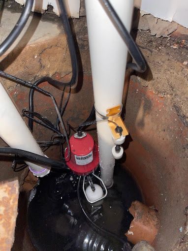 Sump pump in a concrete pit with water, black and red pump, white PVC pipes, and float switch.