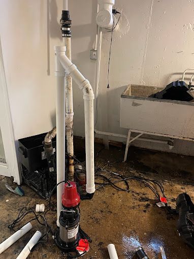 Sump pump installation in a basement. White pipes and black pump on concrete floor with debris.