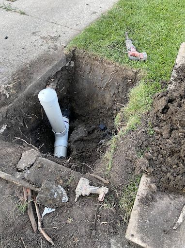 Hole dug in the ground near a sidewalk, revealing a gray pipe for plumbing. Tools and dirt are present.