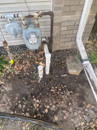 Gas meter, pipes, and white PVC pipes rising from the soil near a house exterior and a gutter downspout.