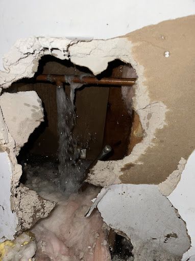 Water gushing from a damaged copper pipe in a hole in a wall, drywall and insulation visible.