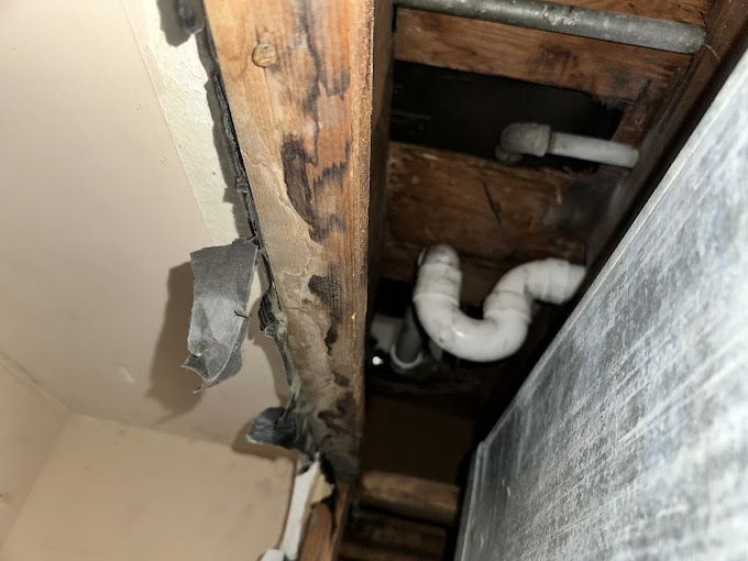 Wood beam with peeling paint, exposing dark stains, and plumbing pipes.