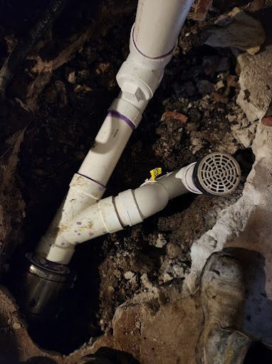 PVC pipes in a dirt hole. A drain assembly is attached. Dark dirt and concrete surround the pipes.