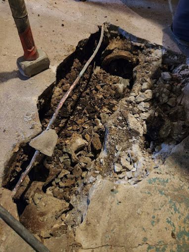 Hole in concrete floor with debris, revealing pipes and a cable. Sledgehammer visible.