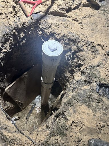 A white pipe extends from a hole in the ground, surrounded by dirt and tools.