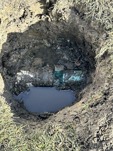 Hole in ground with water, containing a rectangular, turquoise-colored object. Surrounded by dirt and grass.