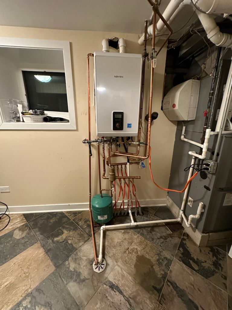 A wall-mounted boiler with copper pipes, a green expansion tank, and white pipes in a basement.
