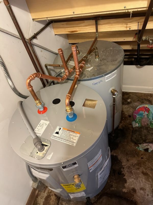 Two water heaters with copper pipes, installed in a damp, unfinished space.