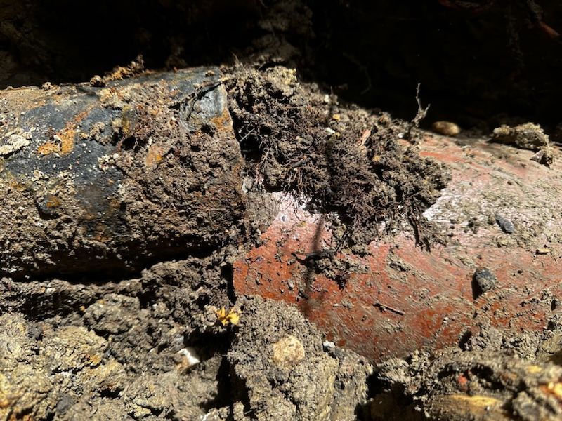 Close-up of a damaged pipe in soil, showing a black section and a reddish-brown section, surrounded by dirt.