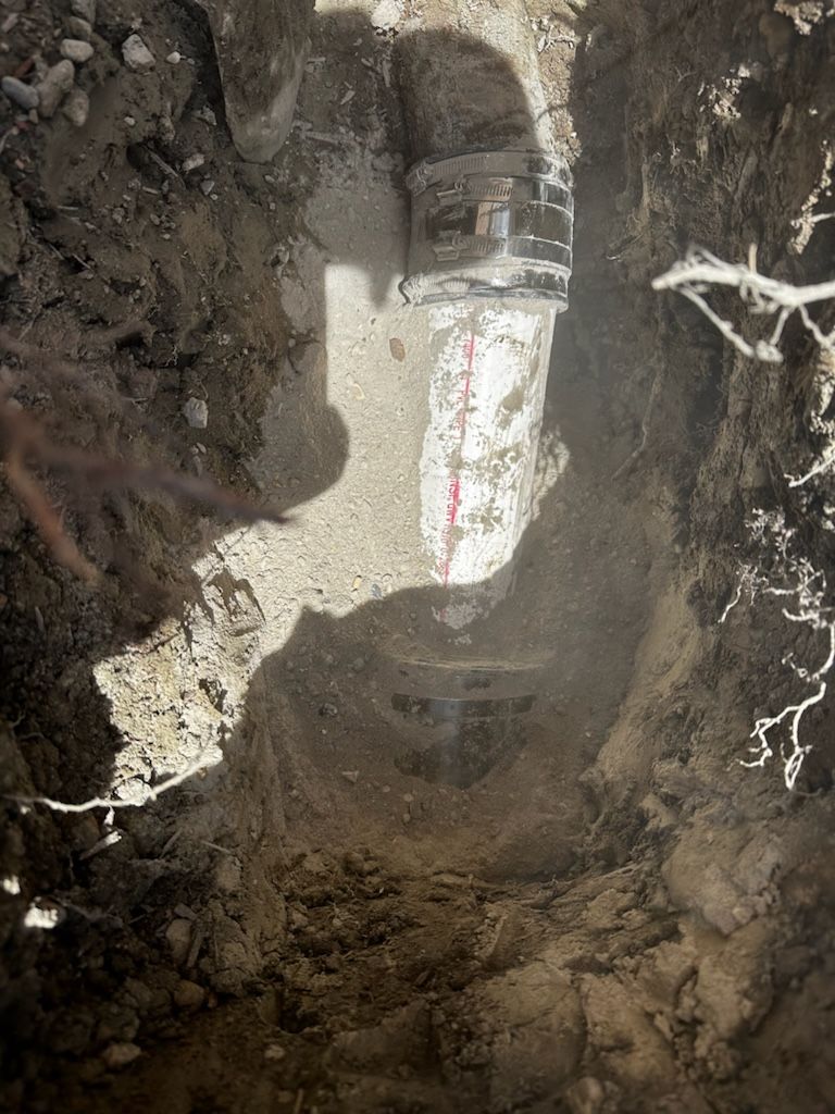 Trench dug around a cylindrical object. Soil surrounds. Bright overhead light. Red and white measuring tape visible.