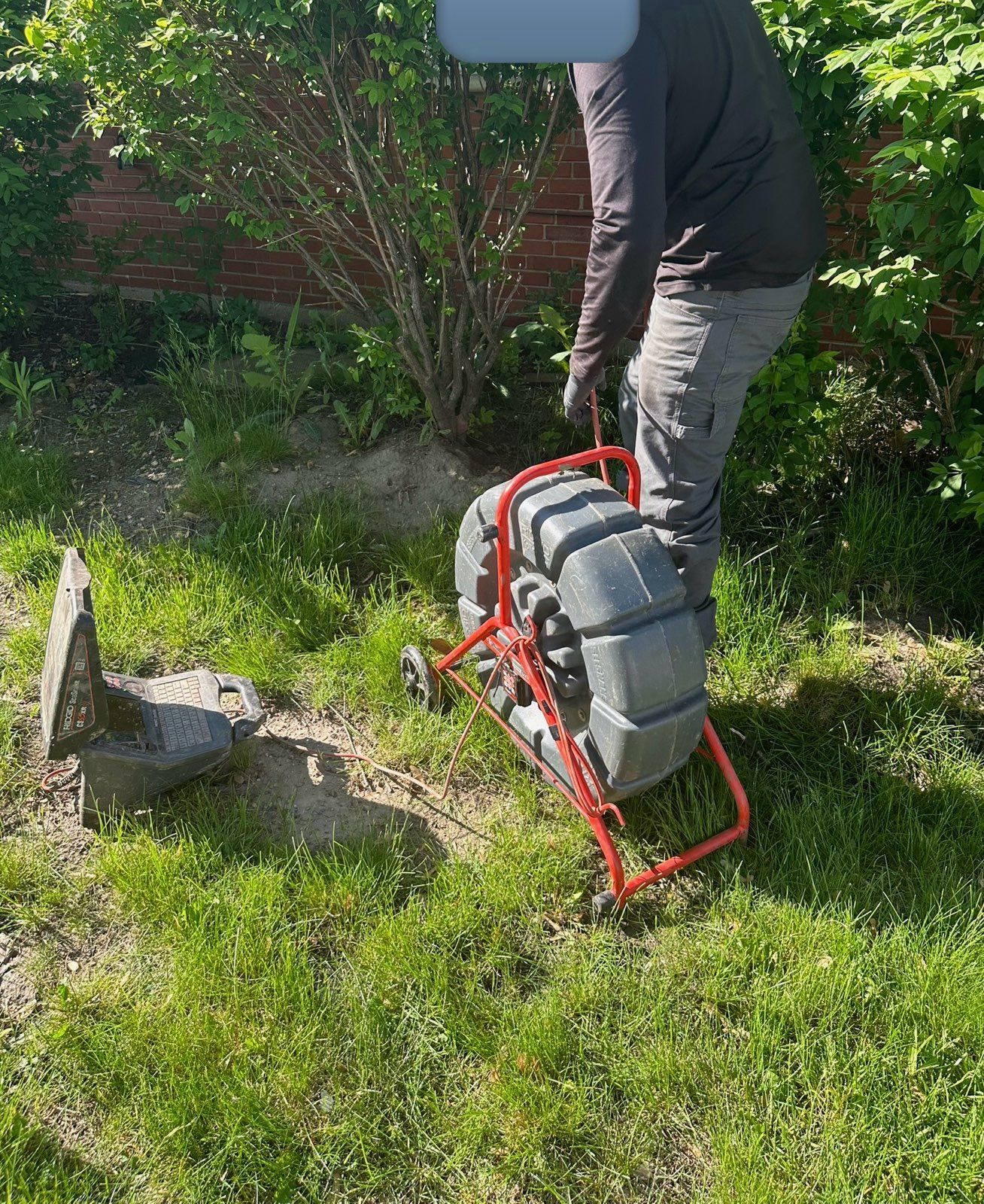 Person lifting a red frame with a large, coiled, gray cable over grass next to a concrete cover.