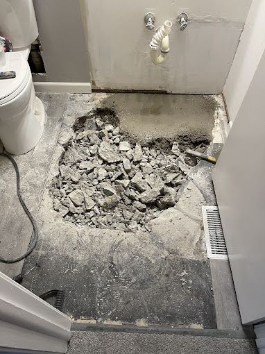 Bathroom floor torn out, exposing concrete rubble and underlayment. Toilet and sink visible in the room.