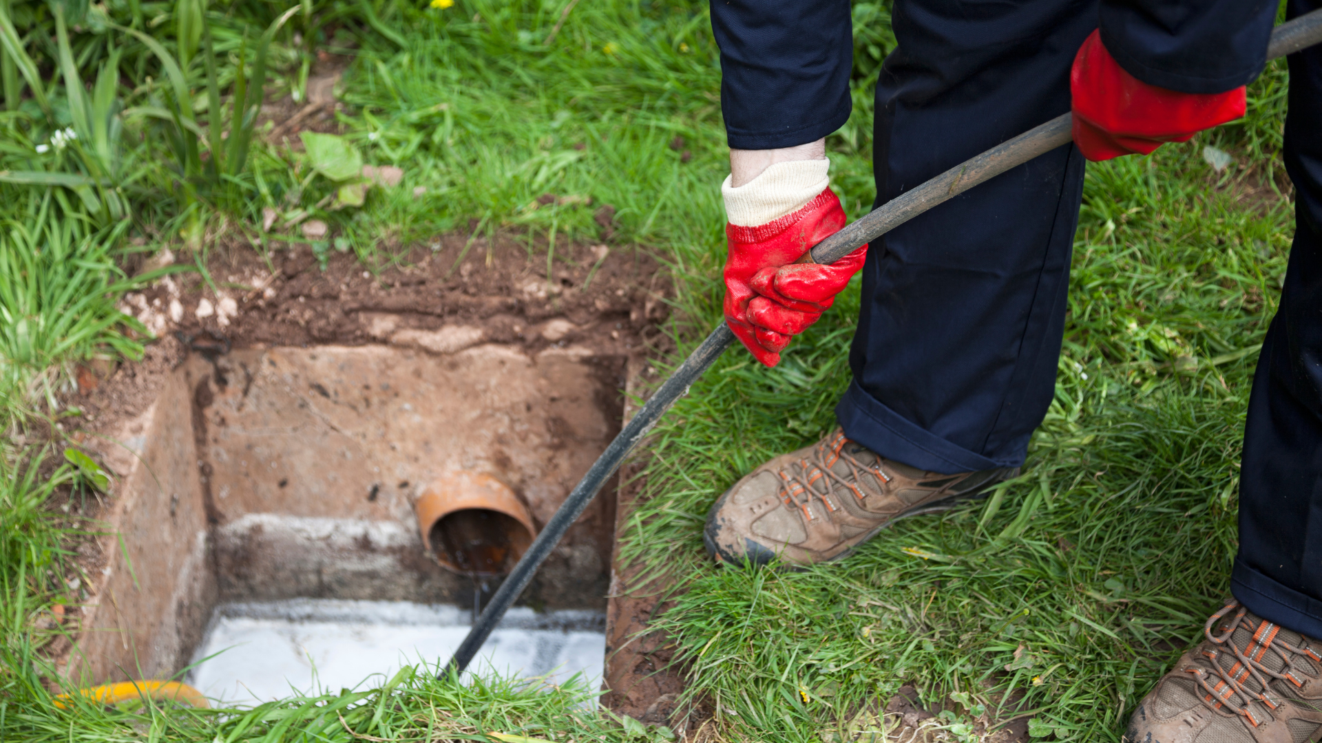 Person in red gloves using a drain snake on an open underground drain in a grassy area.