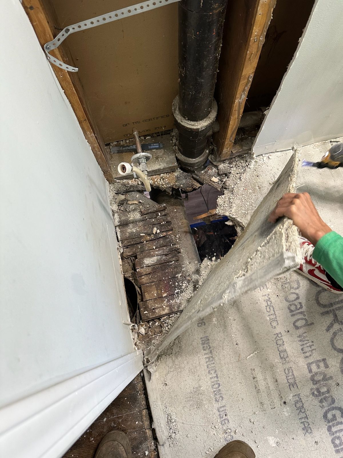 Exposed plumbing and damaged floorboards in a construction area, person holding a piece of drywall.