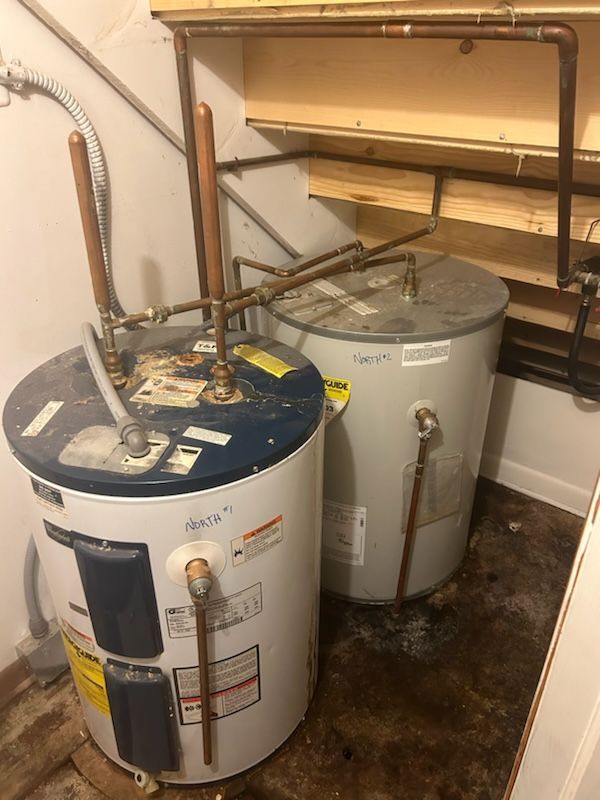 Two water heaters side-by-side in a utility room. Copper pipes connect them to a shelf and wall.