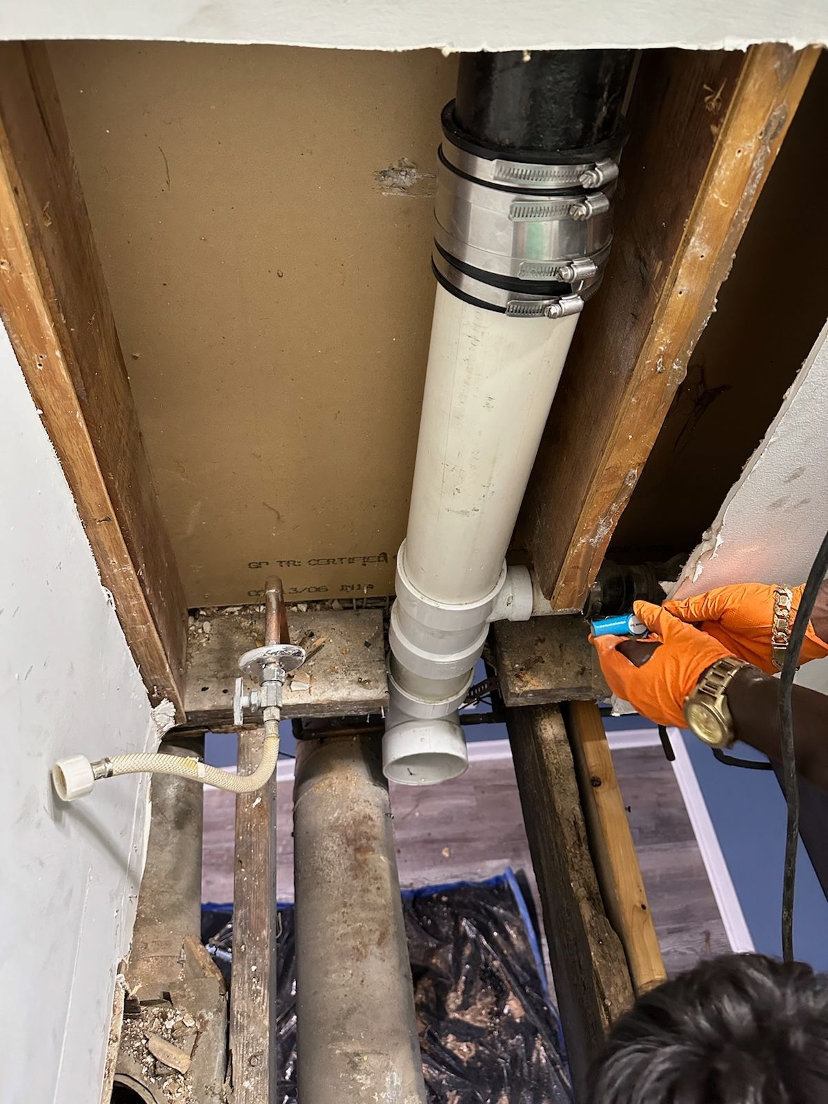 Plumbing work: White and black pipes connected under a ceiling, with orange-gloved hands working.