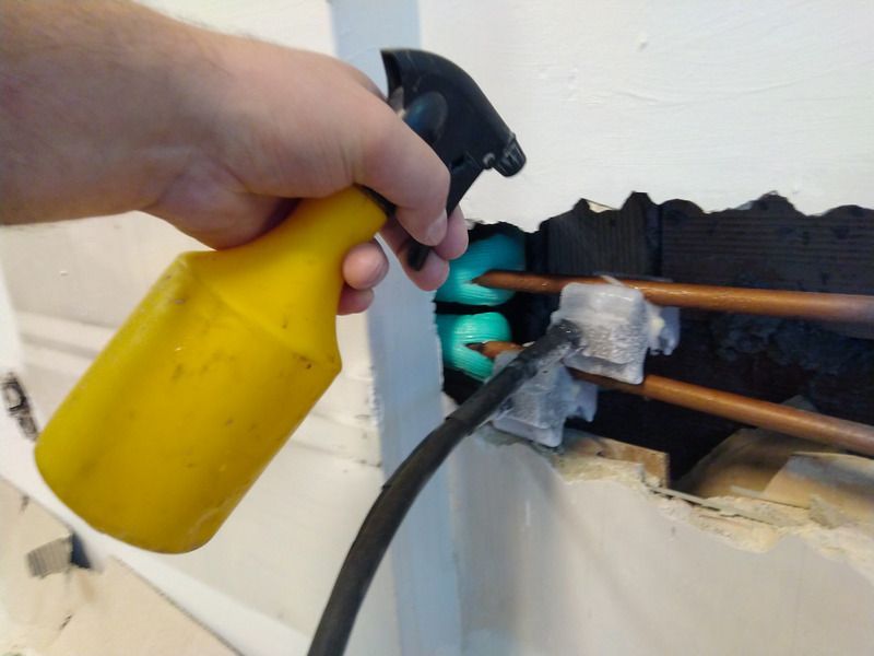 Hand spraying a glowing foam insulator around copper pipes coming out of a wall.