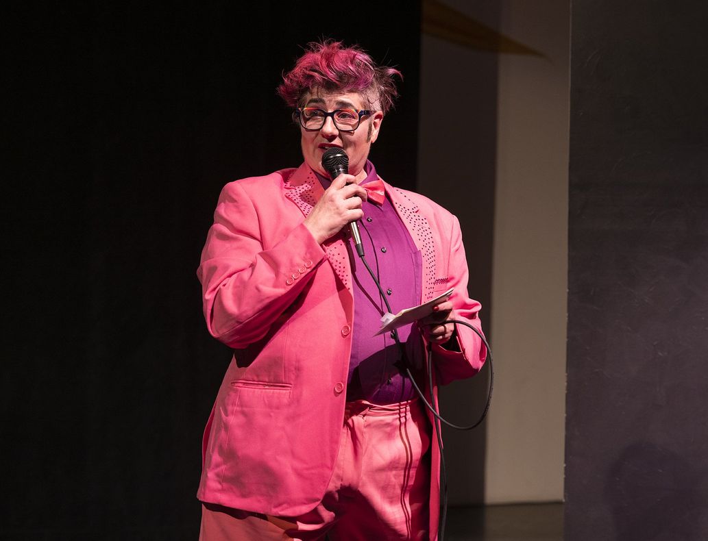 Adam Bomb, Quiver and Tempt Society's EmCee , is holding a mic and looking to the audience while wearing a pink two piece suit and a purple button down shirt.