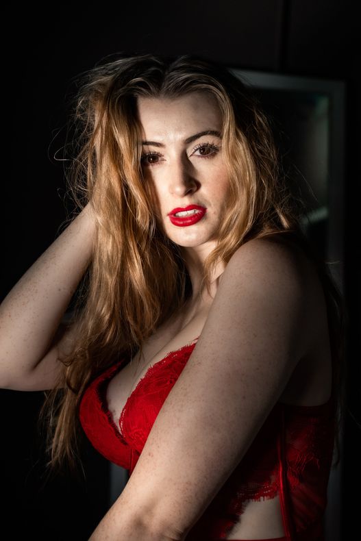 Farleigh Rey, Quiver and Tempt Society performer, is scrunching her hair with her right hand while looking sultry in a red lacy bra and red lipstick.