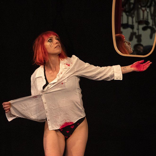 Freya La Fierce, A Quiver and Tempt Society performer, is on stage holding a large mirror, looking off into the distance, and  wearing a large white button down shirt and panties with hearts on them with blood splatters.