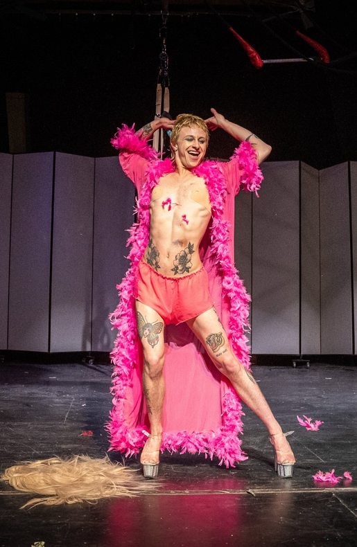 They Von Gay, Quiver and Tempt Society Performer posing on stage wearing a pink feather boa, red workout shorts and clear, platform heals.