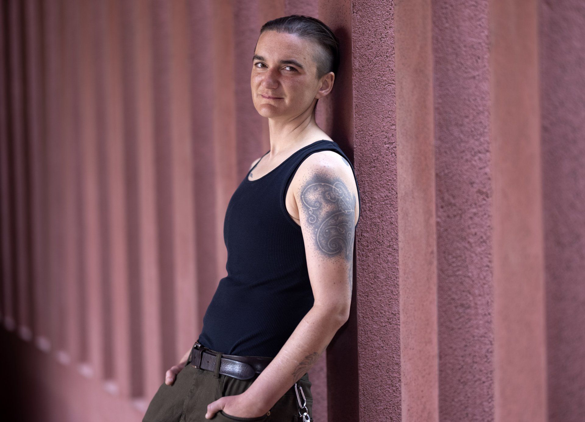 kim g standing against a terracotta colored concrete wall. They are wearing a black tank top with green pants, a swirling tattoo covering their left shoulder.